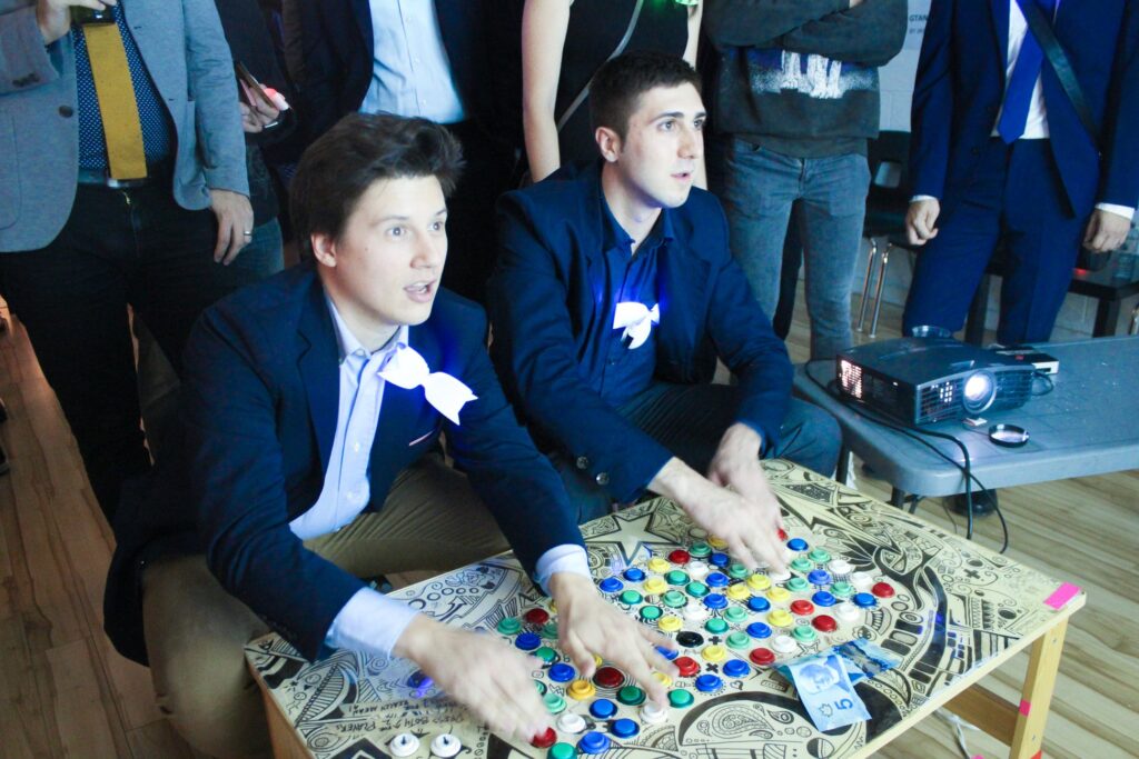 Two sharply dressed men playing a 100 button fight stick. There is Canadian money on the table between them.
