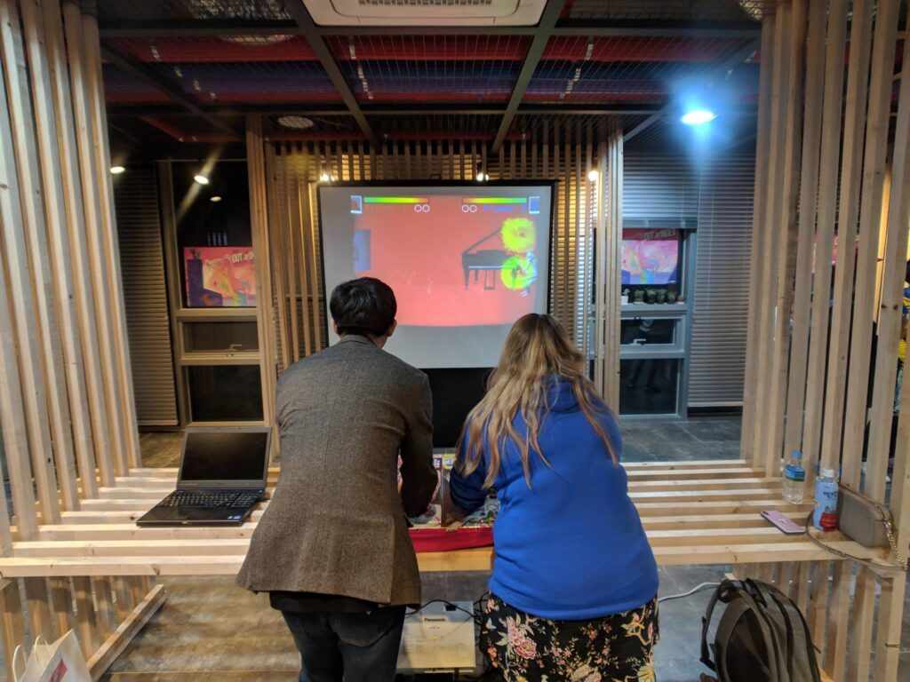 An over the shoulder shot of a man and a woman playing with something out of sight. There is a screen in front of them showing a fighting game. They are surrounded by a wooden frame.