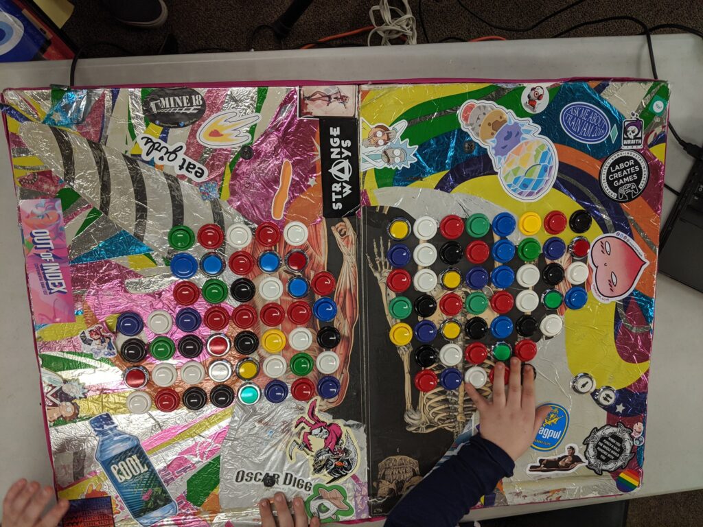 A top down view of a strange controller with 100 buttons. The board itself is covered in stickers and a collage made out of paper and a unicorn balloon.