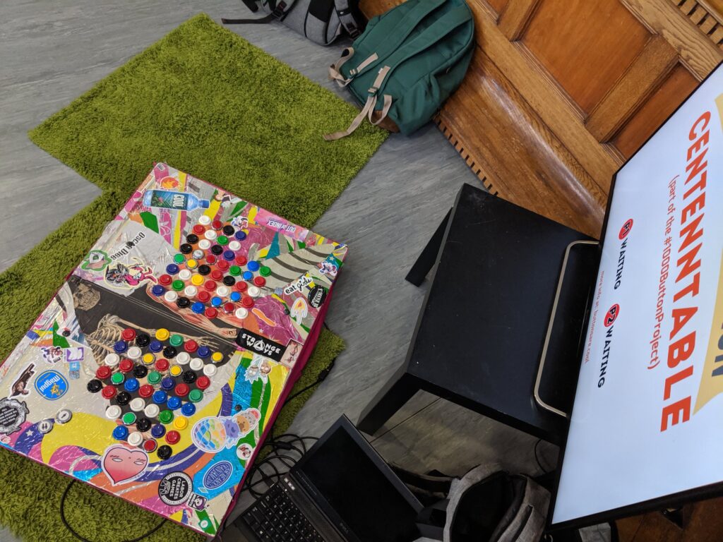 A distance shot of a set up of a elaborate and brightly colored board with 100 buttons and the TV it is attached to that says Centenntable.