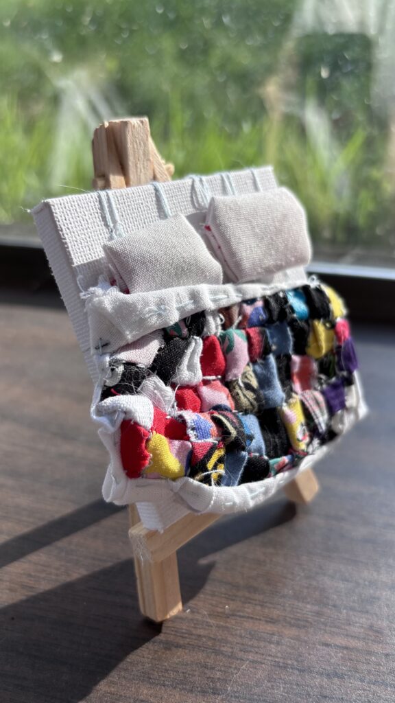 On a wooden surface a small canvas with a miniature quilt in several rainbow squares, and two tiny pillows.