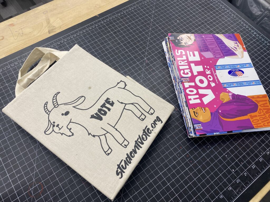 On a self healing mat, a tab bound book made with a poster cover and a book box made out of a tote bag with a goat on it.