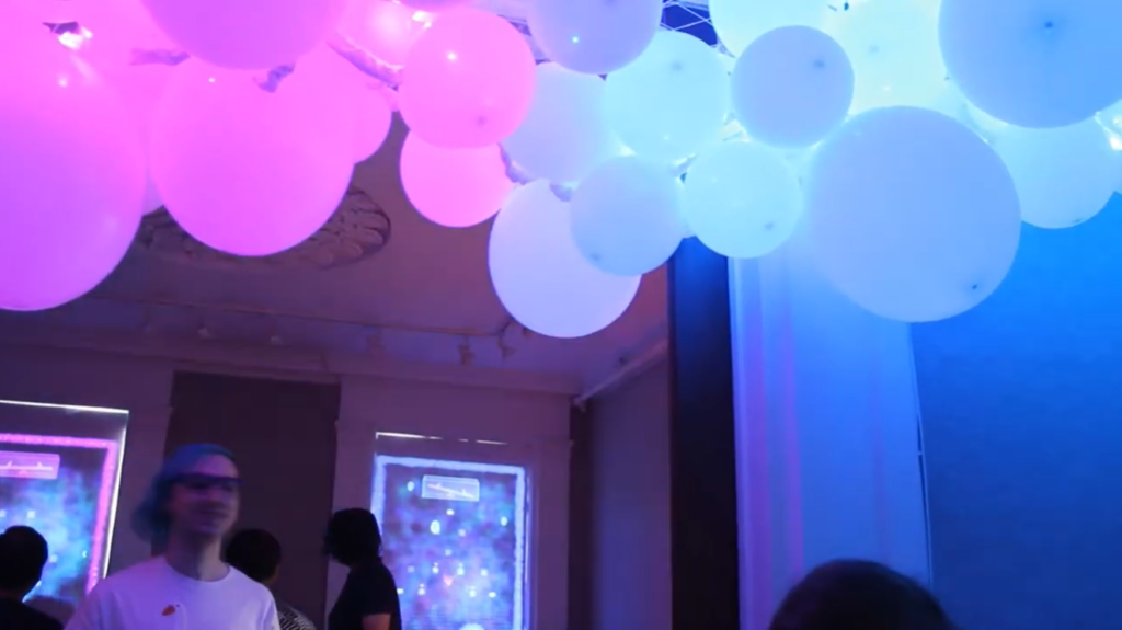 In a crowded gallery, a large ceiling installation made out of blue and purple lit balloons. The room is otherwise dark.